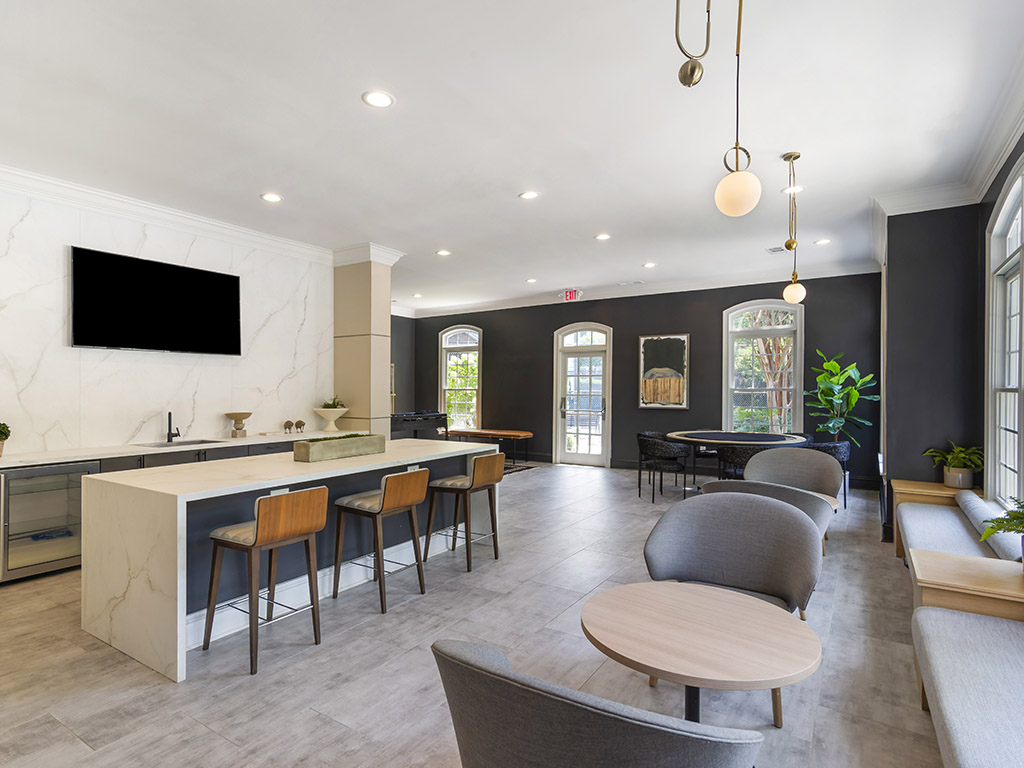 Modern lounge with gray chairs, round tables, a marble island with wooden barstools, large windows, dark accent walls, hanging lights, a large TV, and green plants near the entrance. Natural light fills the space.