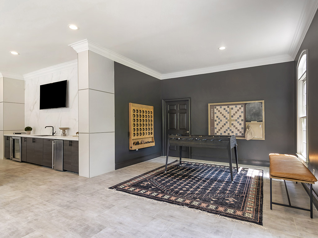 Modern game room with a foosball table on a patterned rug, wall games, a bench, a TV above a sink, and cabinets against gray and white walls with large windows letting in natural light.