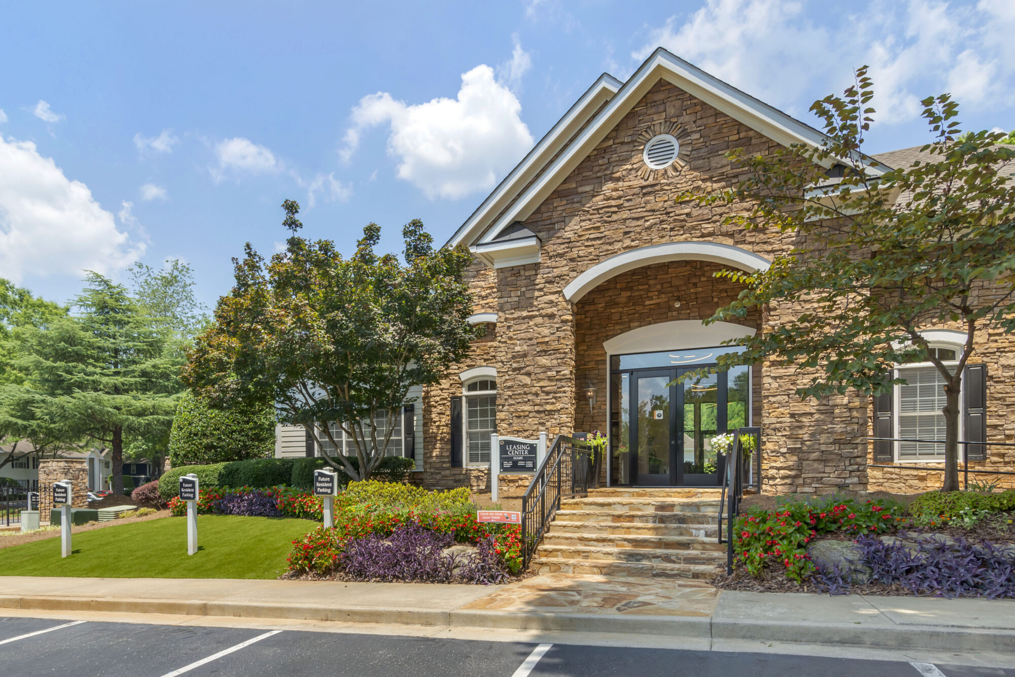 A stone building with white trim, landscaped yard, and stairs leading to a glass door. Signs and greenery surround the entrance under a blue sky with clouds.