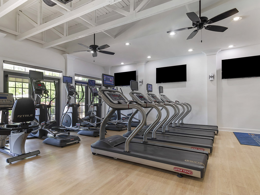 A modern gym with treadmills, stationary bikes, large windows, several wall-mounted TVs, ceiling fans, and light wood floors. The room is well-lit and spacious, with exercise equipment arranged neatly.