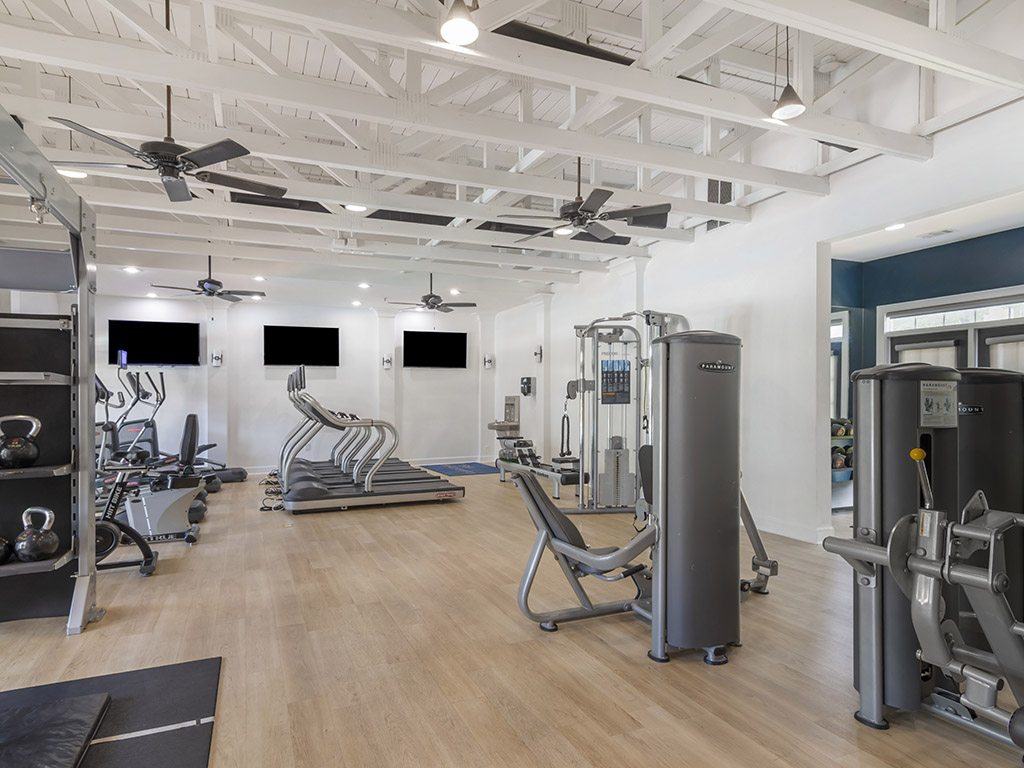 Modern gym with wooden floors, various exercise machines, treadmills, elliptical trainers, kettlebells, and large windows. White ceiling beams and ceiling fans are visible, creating a bright and spacious workout environment.