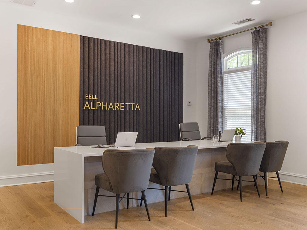 Modern office with a white desk, four gray chairs, two laptops, and wall art displaying Bell Alpharetta on wood and dark panels. Large window with gray curtains lets in natural light.