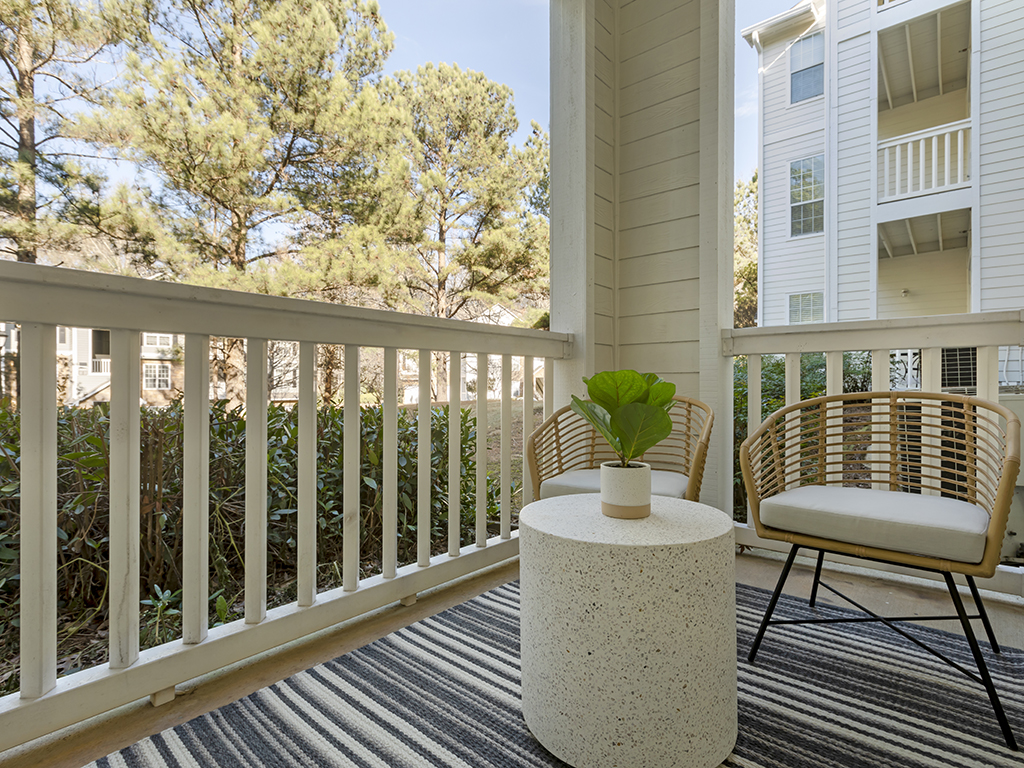 A cozy balcony features two wicker chairs, a round white table with a potted plant, and a striped rug. Trees and parts of a white building are visible in the background.