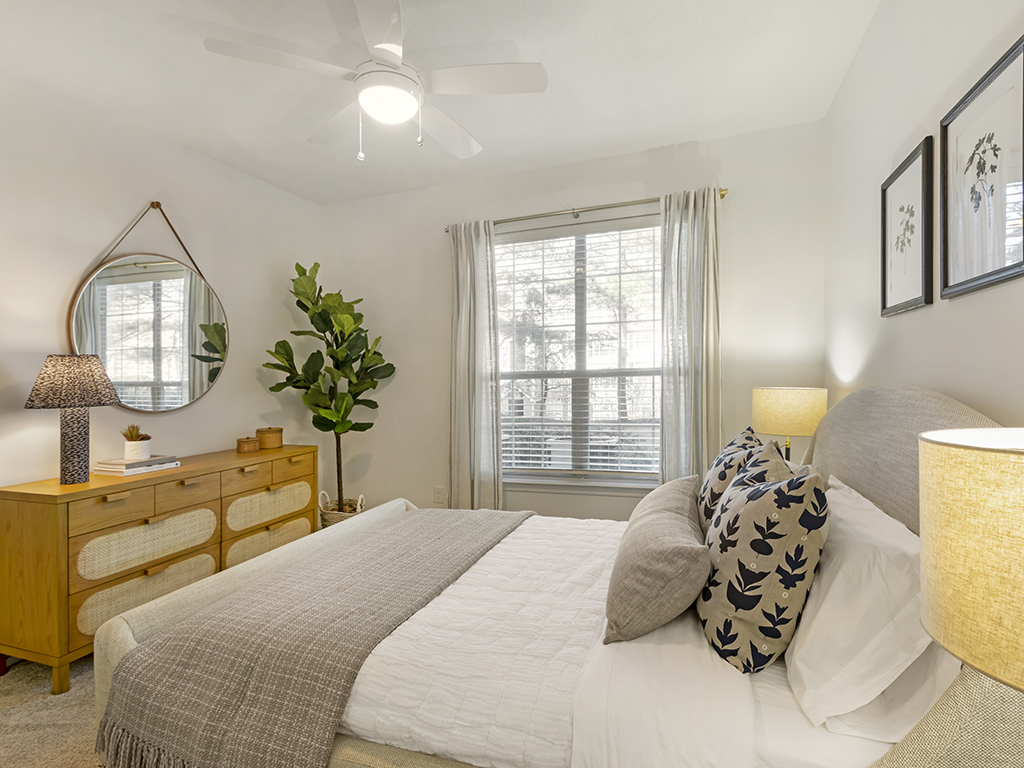 A cozy bedroom with a large bed, neutral bedding, wooden dresser, indoor plant, lamps, wall art, and a window letting in natural light. The decor is modern and inviting.