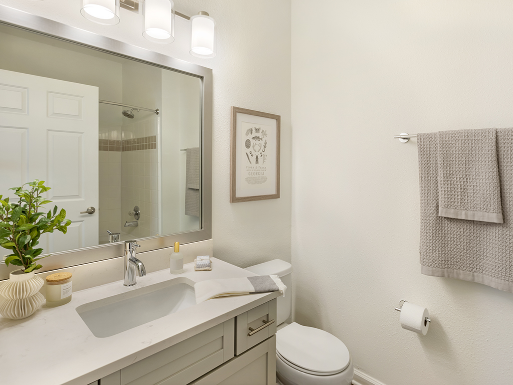 Modern bathroom with a white sink, light gray cabinets, a mirror, a plant, towel rack with gray towels, wall art, toilet, and beige walls. Soft lighting from three lights above the mirror.