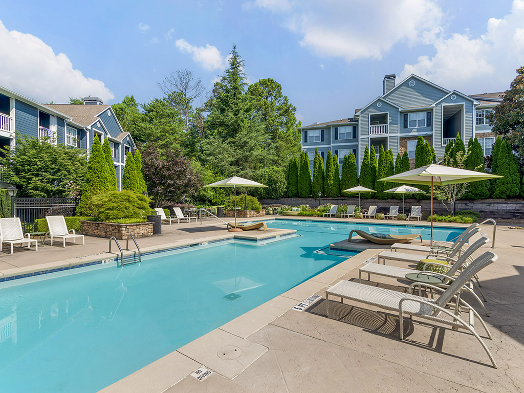 Outdoor swimming pool with lounge chairs, umbrellas, and landscaped greenery, set in front of blue apartment buildings on a sunny day.