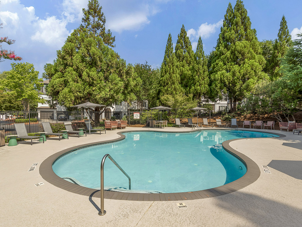 Outdoor swimming pool with a curved shape, surrounded by lounge chairs, umbrellas, and trees. The pool area is empty and the sky above is partly cloudy.