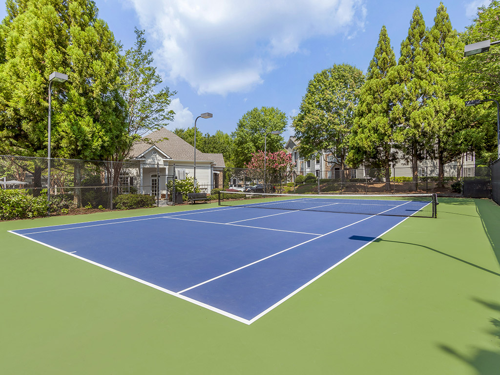 A blue and green outdoor tennis court is surrounded by trees and residential houses under a partly cloudy sky.