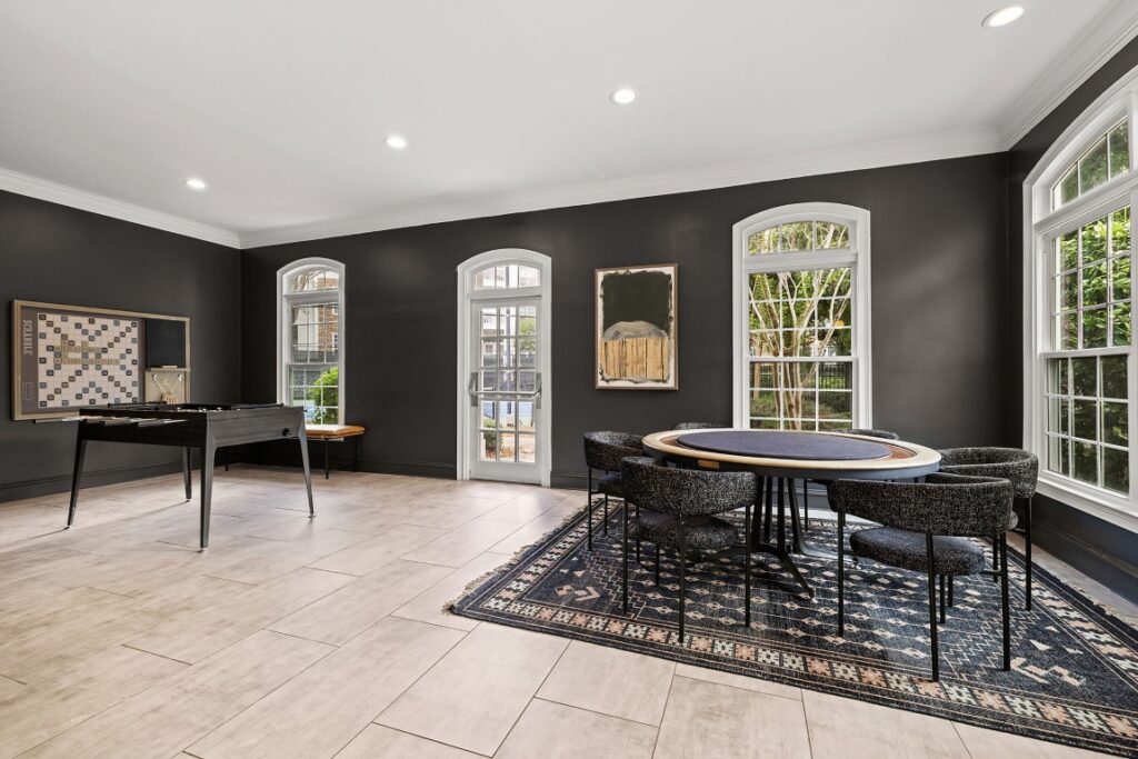 A game room with charcoal walls featuring a large wall-mounted Scrabble board, a foosball table, and a circular game table with six chairs atop a patterned area rug.