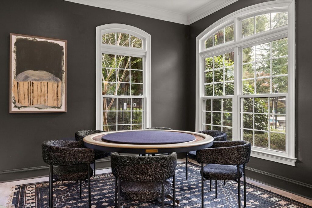 A circular game table with a purple felt top surrounded by four modern chairs in a room with dark gray walls and two large windows.
