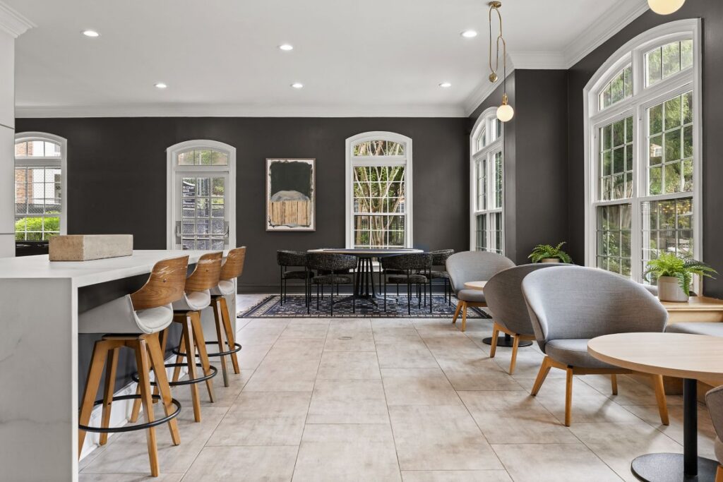Modern lounge area with a kitchen island featuring wooden bar stools, a dining table with chairs, and a seating area with grey armchairs and small round tables.