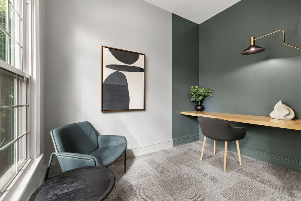 A small home office featuring a floating wooden desk, a dark grey chair, a teal armchair, and abstract wall art, set against light grey and dark green walls.