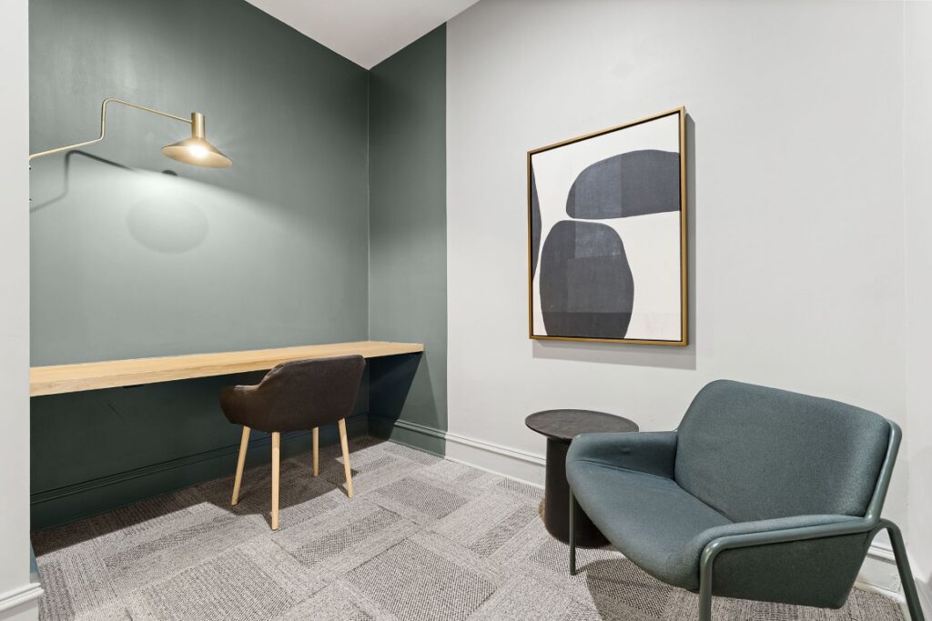 A home office corner with a built-in wall desk, a dark chair, a small side table, an armchair, and an abstract black-and-white wall art piece.