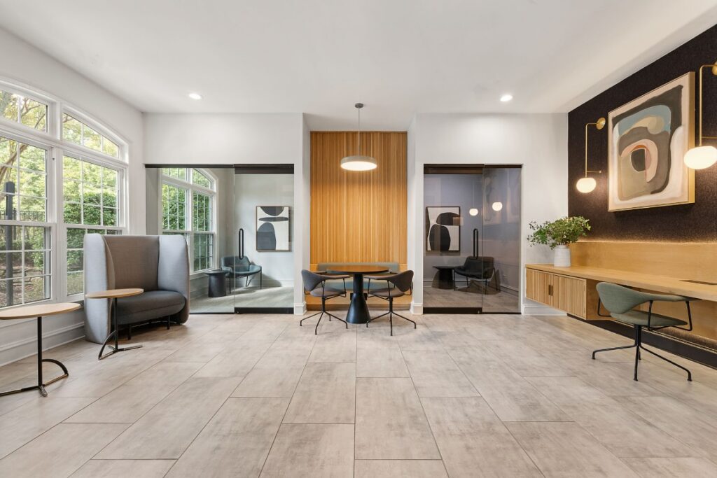 A modern lobby or coworking area featuring a central table with chairs, a built-in desk with seating, a high-backed lounge chair, and two glass-enclosed private offices.