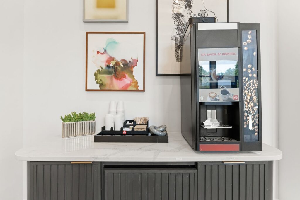 A black coffee machine sits on a white countertop next to a tray of coffee cups, stirrers, and sweeteners, with abstract framed art on the wall behind it.