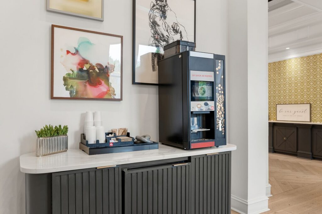 An office coffee machine on a marble countertop with a stack of paper cups, wooden stirrers, and a small potted plant, framed by artwork on the wall.