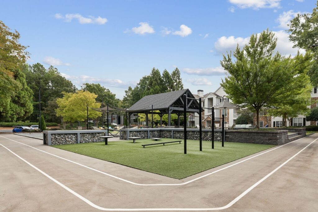 Outdoor recreational area with a central pavilion, fitness equipment, and low stone walls on a turf surface, surrounded by a paved walking path.