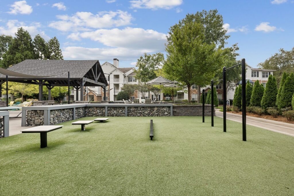 Outdoor fitness area with exercise equipment and a covered pavilion on a lawn in front of apartment buildings.
