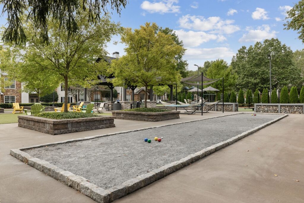 A bocce ball court with three balls on the gravel, surrounded by trees, raised garden beds, and outdoor lounge seating.