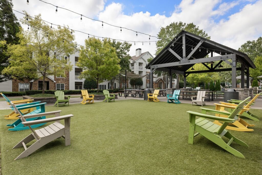 Colorful Adirondack chairs arranged in a circle on a grassy courtyard, with picnic tables and a wooden pavilion in the background.