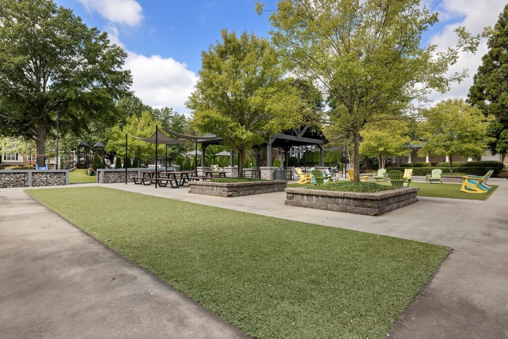 A park courtyard with a paved walking path, artificial turf, picnic tables under a shade sail, and colorful chairs around raised planters.