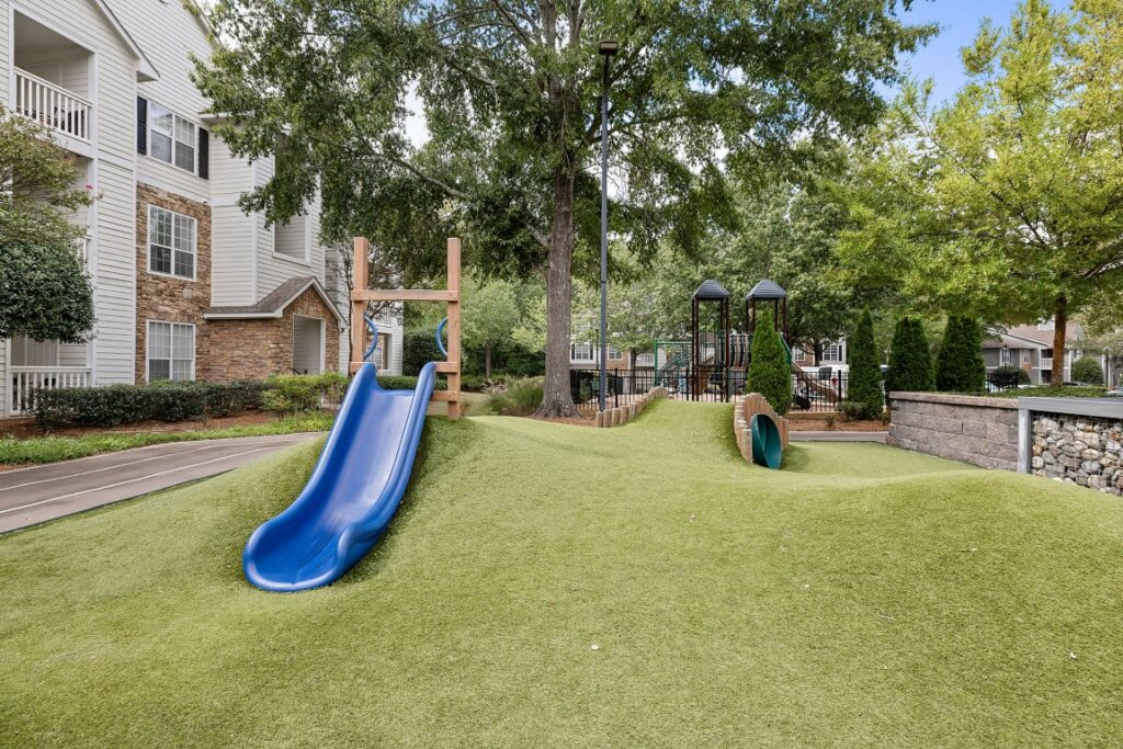 A playground with a blue slide on a grassy hill next to an apartment building.