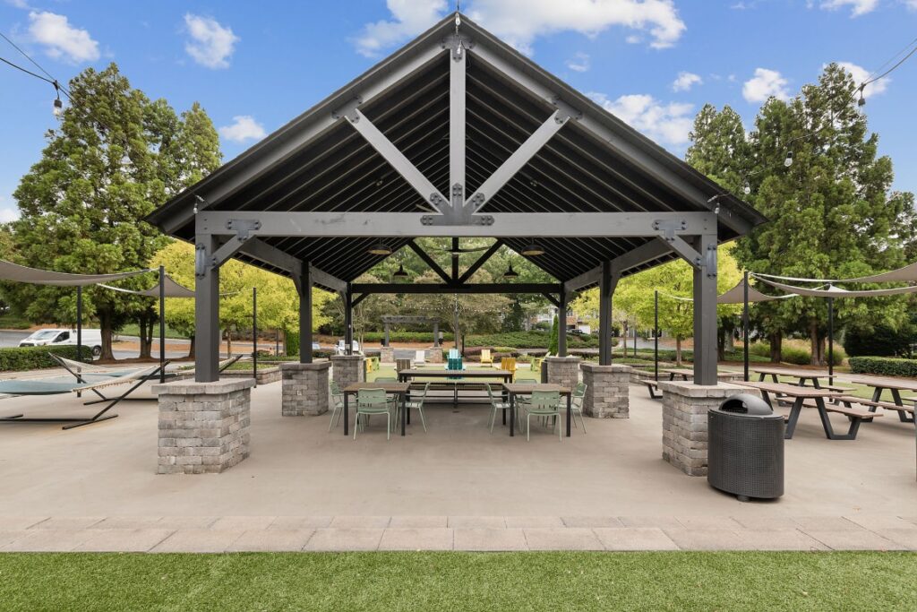An open-air pavilion with a dark metal roof supported by stone pillars, featuring outdoor dining tables, chairs, and a hammock in a park setting.