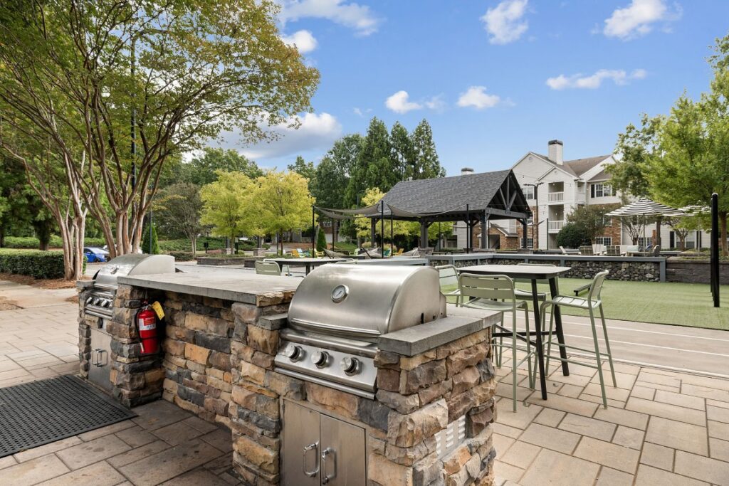 Outdoor kitchen area with a built-in stainless steel grill, stone base, and nearby patio dining table on a paved plaza.