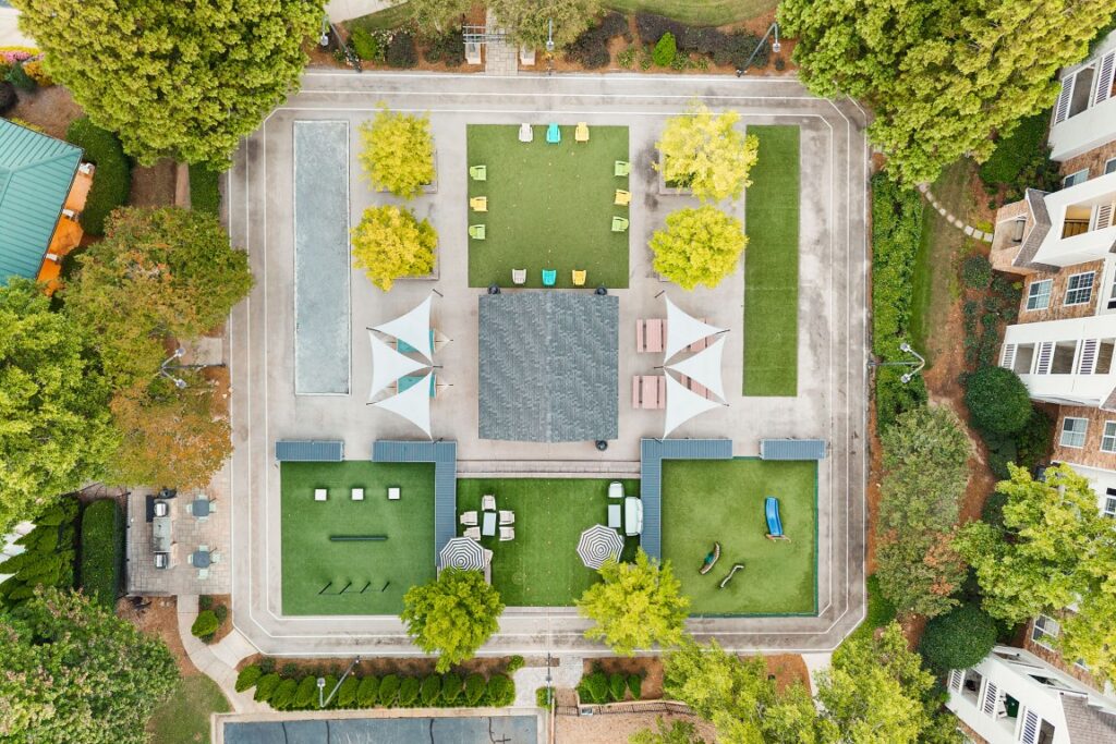Aerial view of a courtyard featuring a central pavilion, artificial turf areas with scattered chairs, picnic tables, and a small playground, all surrounded by trees and residential buildings.