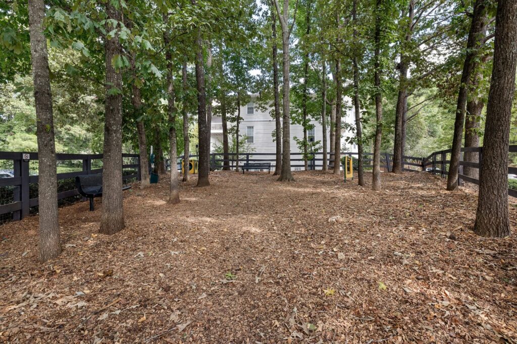 A fenced dog park area covered in wood chips with trees, several benches, and agility equipment.