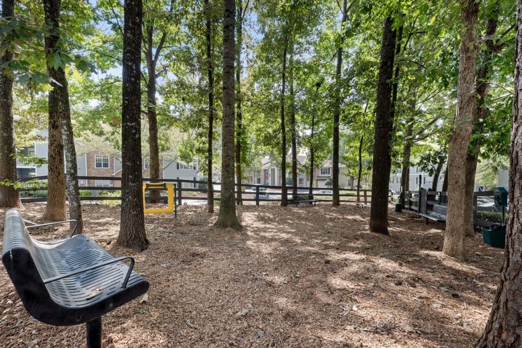 A dog park with wood chip ground cover, several benches, and a yellow agility hoop, surrounded by tall trees and a wooden fence with apartment buildings in the background.