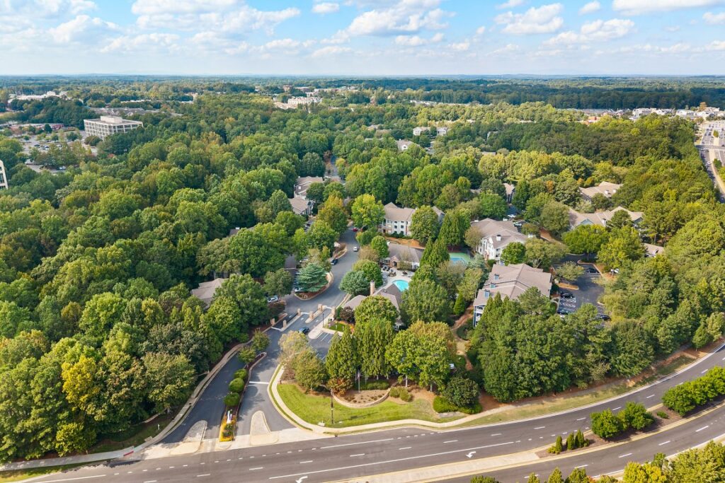 Aerial view of an apartment complex nestled within a dense forest, adjacent to a multi-lane road.