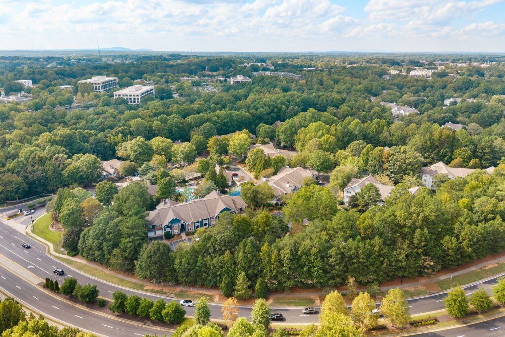 An aerial view of a multi-building apartment complex nestled within a dense forest next to a curved multi-lane road.