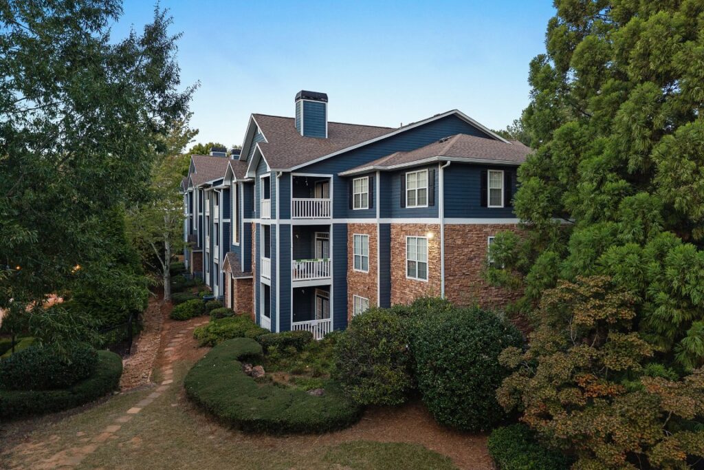 A multi-story blue apartment building with stone siding and balconies surrounded by trees.