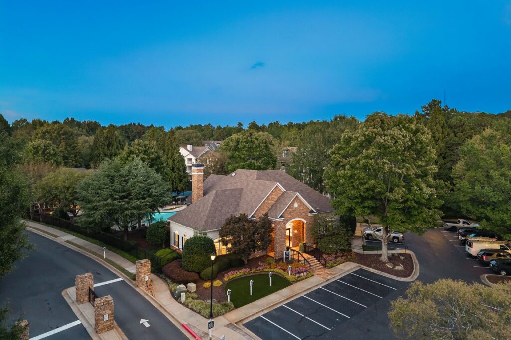 Aerial view of a brick clubhouse with a peaked roof, adjacent parking lot, and pool, surrounded by trees at dusk.