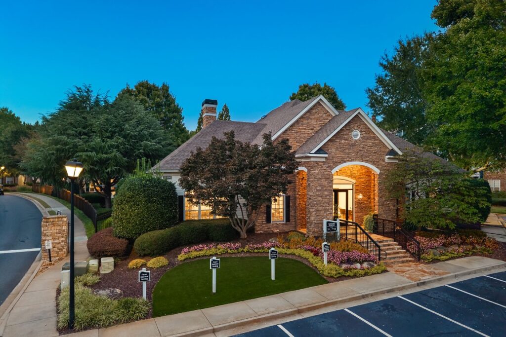 An aerial view of a stone-faced building at dusk, featuring a manicured lawn with parking signs, a front entrance with steps, and an adjacent parking lot.