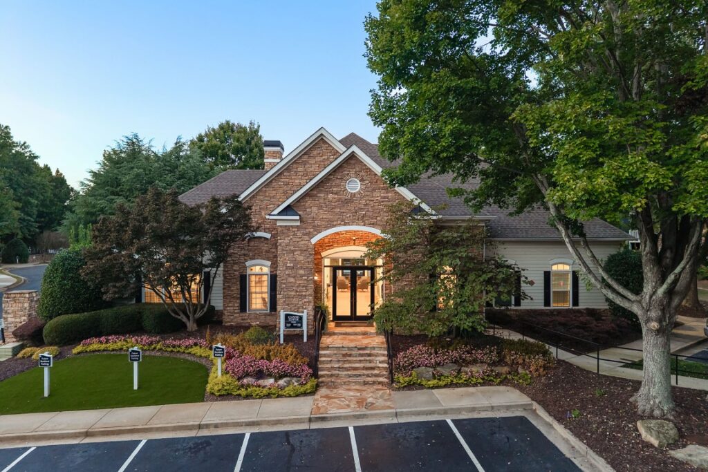 A stone-front apartment leasing office with a front entrance staircase, surrounding landscaping, and a parking lot in the foreground.