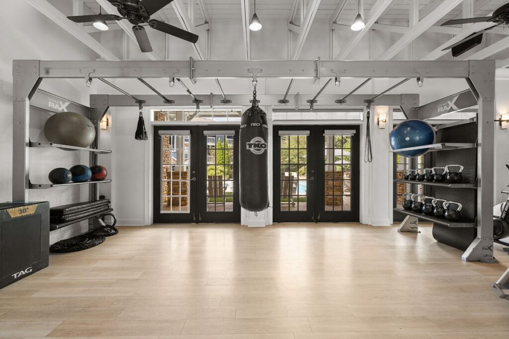 A home gym featuring a central heavy punching bag, wall-mounted storage racks holding medicine balls and kettlebells, and ceiling-mounted monkey bars, all positioned in front of double glass doors.