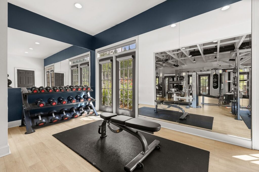 A home gym featuring a weight rack with dumbbells, an adjustable workout bench on a rubber mat, and a large wall mirror reflecting gym equipment.