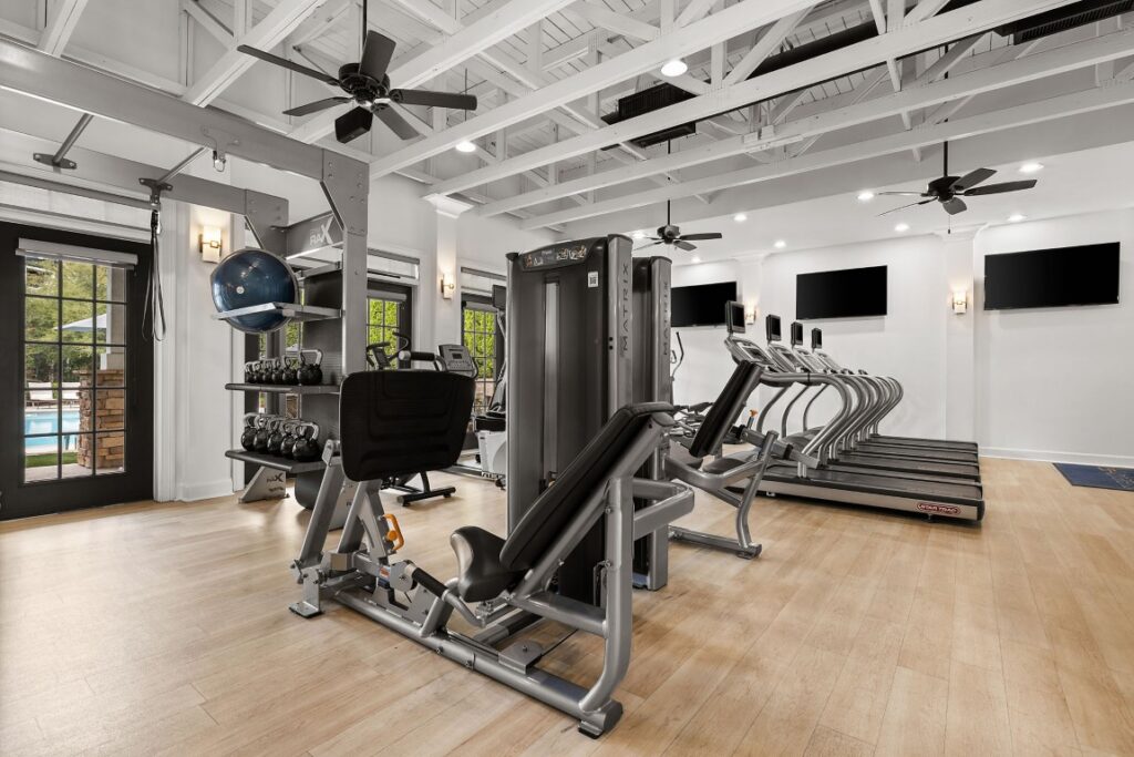 Gym interior with a weight machine, dumbbell rack, and a row of treadmills facing wall-mounted televisions.