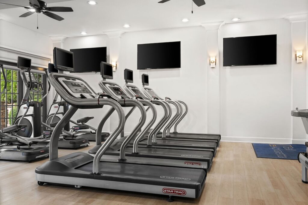 A row of five treadmills in a gym, each equipped with a screen, lined up in front of wall-mounted televisions.