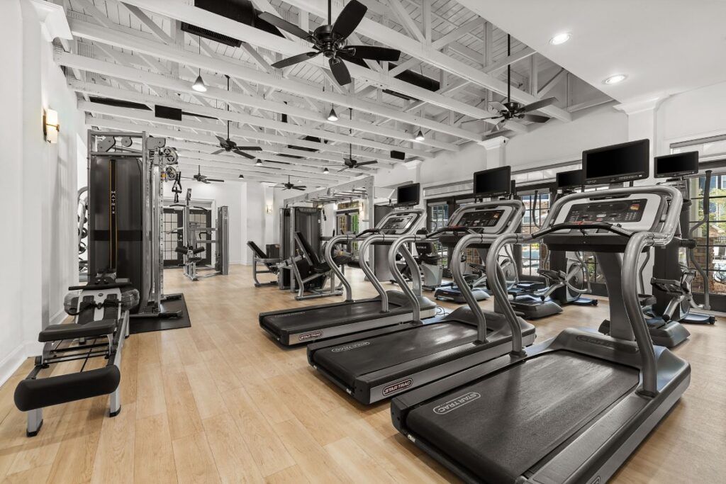 A fitness center with several treadmills, weight machines, and ceiling fans in a room with a white exposed-beam ceiling and light-colored wood flooring.