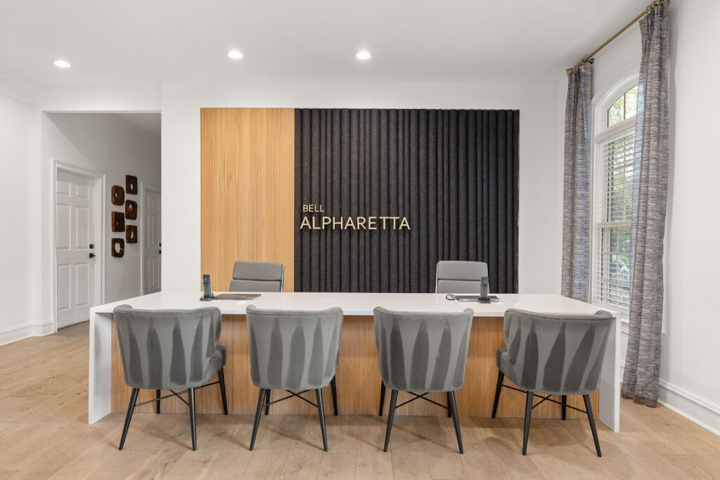 A reception desk featuring the sign Bell Alpharetta on a paneled wall, with four grey upholstered chairs facing the desk and two grey office chairs behind it.