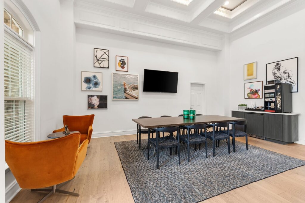 A modern meeting room with a large wooden table, eight black chairs, a wall-mounted television, a gallery wall of framed art, and two orange lounge chairs in the foreground.