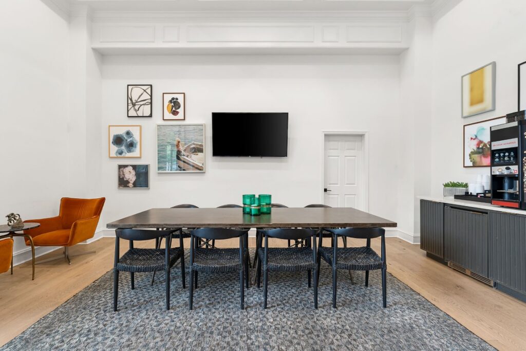 A dining room with a long table, six black chairs, a gallery wall with framed art, a mounted television, and a coffee station.