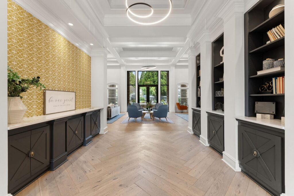 A spacious, symmetrical foyer with herringbone wood floors, white trim, dark storage cabinets, and a yellow patterned accent wall leading to a sunlit seating area with blue armchairs.