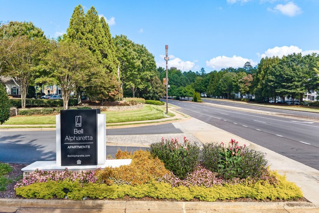 A black and white sign for Bell Alpharetta Apartments situated in a landscaped garden bed next to a multi-lane road.