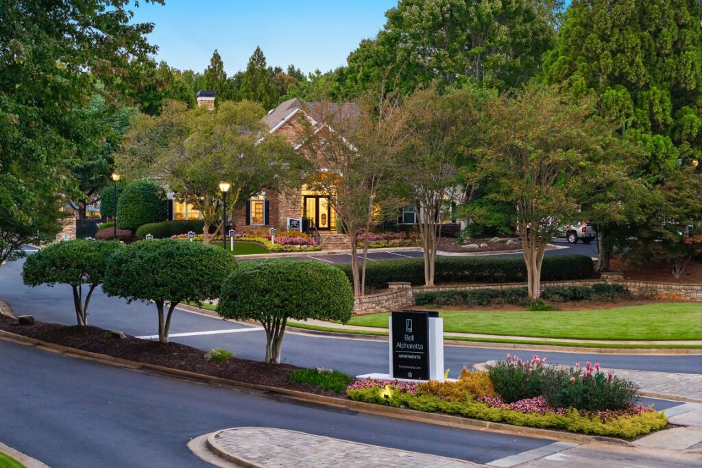 A view of the Bell Alpharetta apartment complex, featuring a brick clubhouse building set behind a landscaped entrance with a monument sign.
