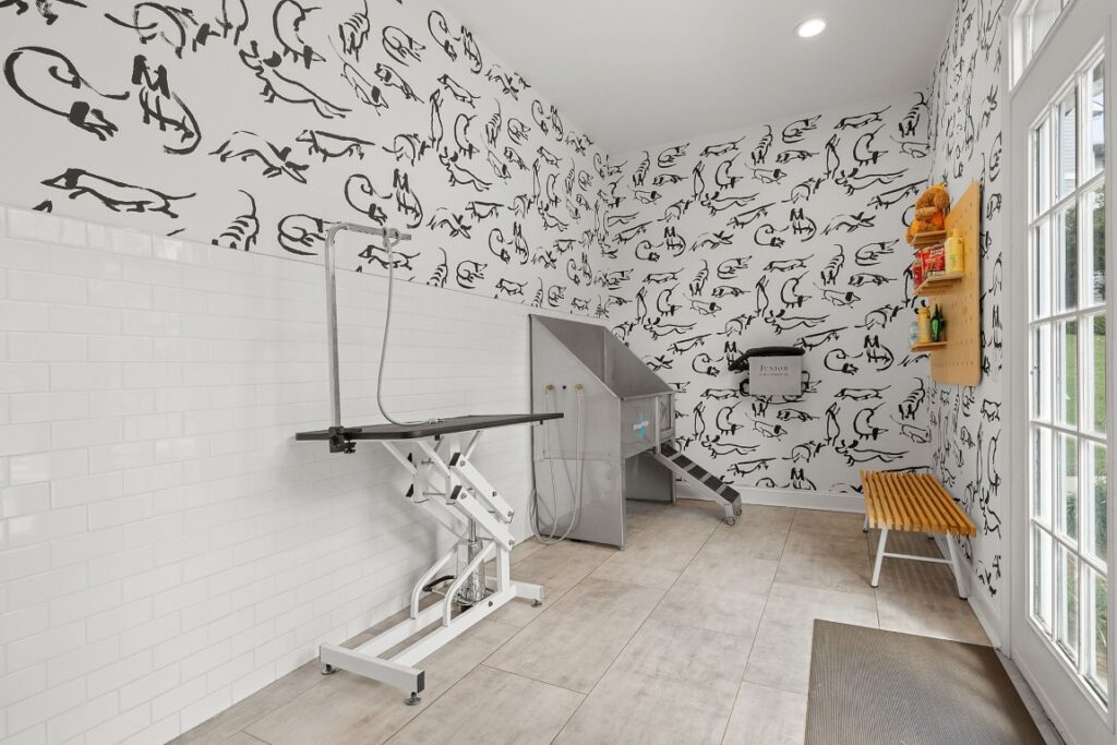 A pet grooming station with an adjustable table, a stainless steel wash tub with steps, and a wooden bench against a wall covered in dog-themed wallpaper.