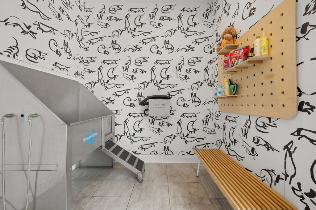 A dog wash station featuring a stainless steel tub with a ramp, a wooden pegboard shelf with pet supplies, and a slatted wood bench against a wall covered in dog-themed wallpaper.